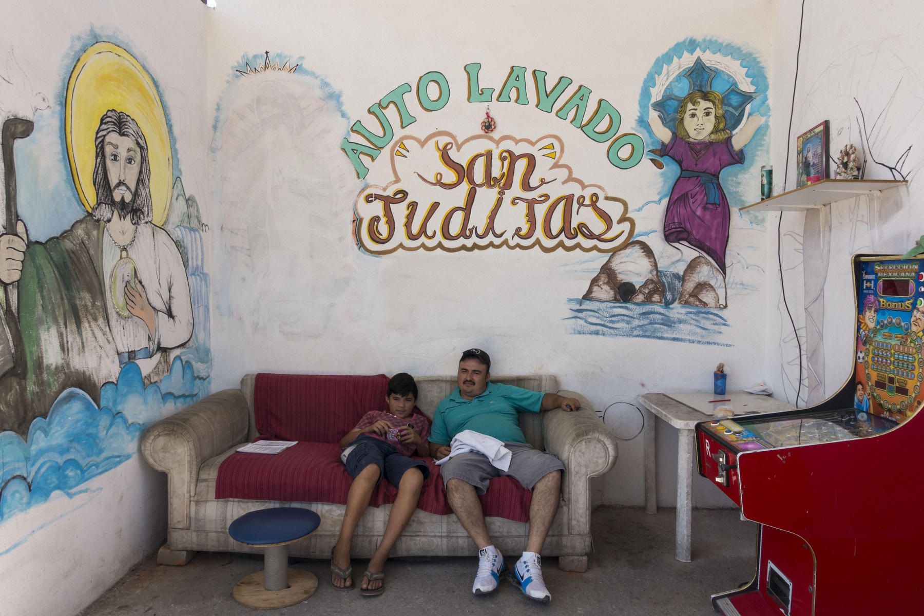 A father and son resting on sofa surrounded by religious murals at a car-wash.
 : PUERTO VALLARTA - Wall Art & Bicycle Tour : Viviane Moos |  Documentary Photographer